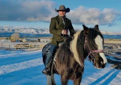A person wearing a cowboy hat and green coat rides a black and white horse with a purple bridle through a snowy rural area. Wooden fences and scattered buildings line the snow-covered ground, with snow-capped mountains in the background under a partly cloudy sky. Long shadows suggest early morning or late afternoon light, adding depth to the wintry landscape.