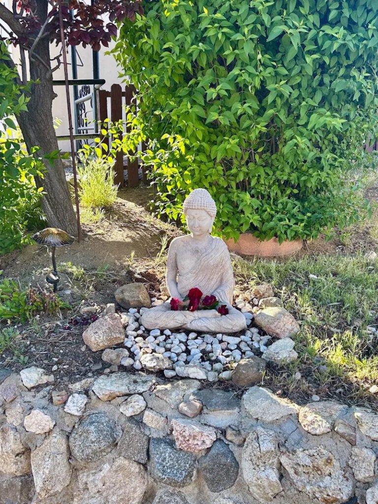 - Garden Buddha Statue with Flowers and Rocks A Buddha statue sits on white pebbles, surrounded by rocks and red flowers. Green foliage and a tree provide a tranquil backdrop, with a garden light nearby.