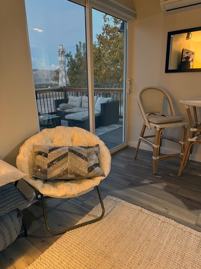 - ndoor Living Space with Patio View A cozy living room featuring a cushioned papasan chair with a chevron pillow, two woven barstools at a counter, and a large sliding glass door opening to a patio with a sectional sofa and autumn trees.