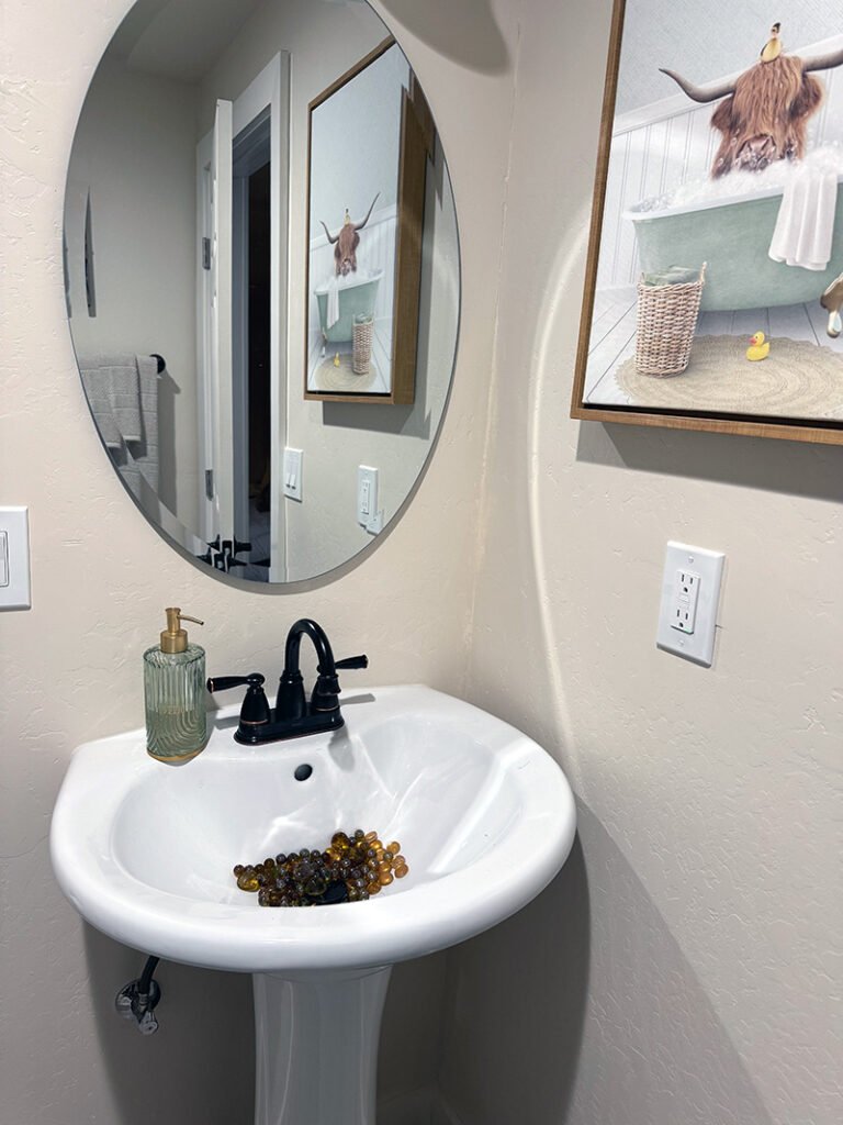 - Bathroom with Highland Cow Artwork A white pedestal sink with a black faucet and ribbed glass soap dispenser sits beneath a round mirror. A framed artwork of a highland cow in a bubble bath adds whimsy to the clean, modern bathroom.