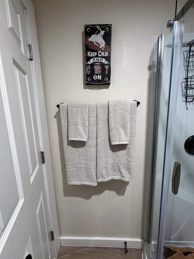 - Western-Themed Bathroom Decor A bathroom wall displays a sign with a cowboy silhouette and the phrase “KEEP CALM AND COWBOY ON” above a towel rack holding three beige towels. A glass shower enclosure is partially visible to the right.