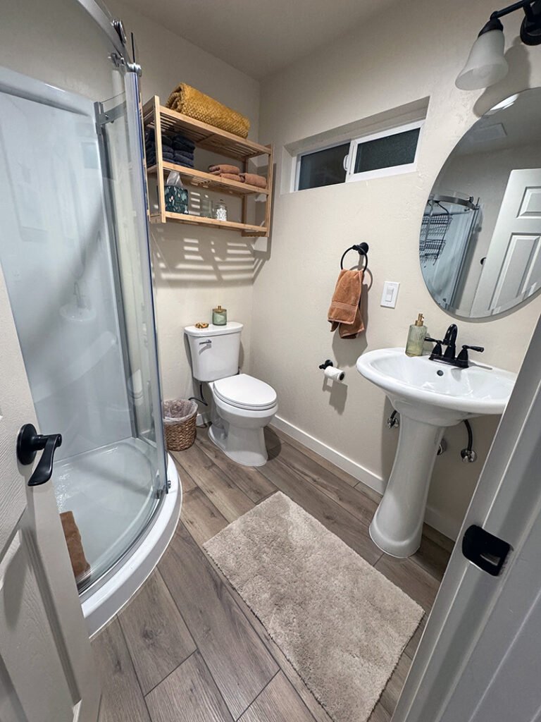 - Compact Bathroom with Corner Shower A modern bathroom with a curved glass shower, pedestal sink, and white toilet. A wooden shelf above the toilet holds folded towels and decor. A round mirror and black towel ring complete the neutral-toned space.