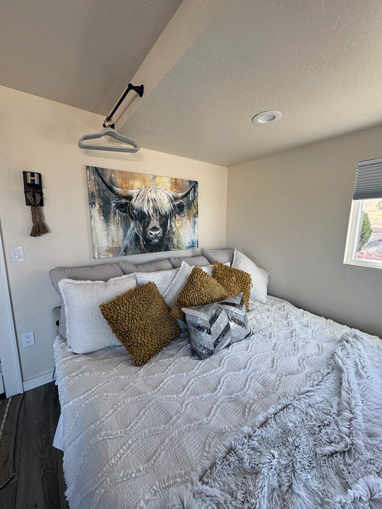 - Bedroom with Highland Cow Painting A neatly made bed with mustard and white pillows sits beneath a large painting of a highland cow. A rope wall accent and ceiling-mounted hanger bar add rustic charm. Natural light enters through a window with blinds.