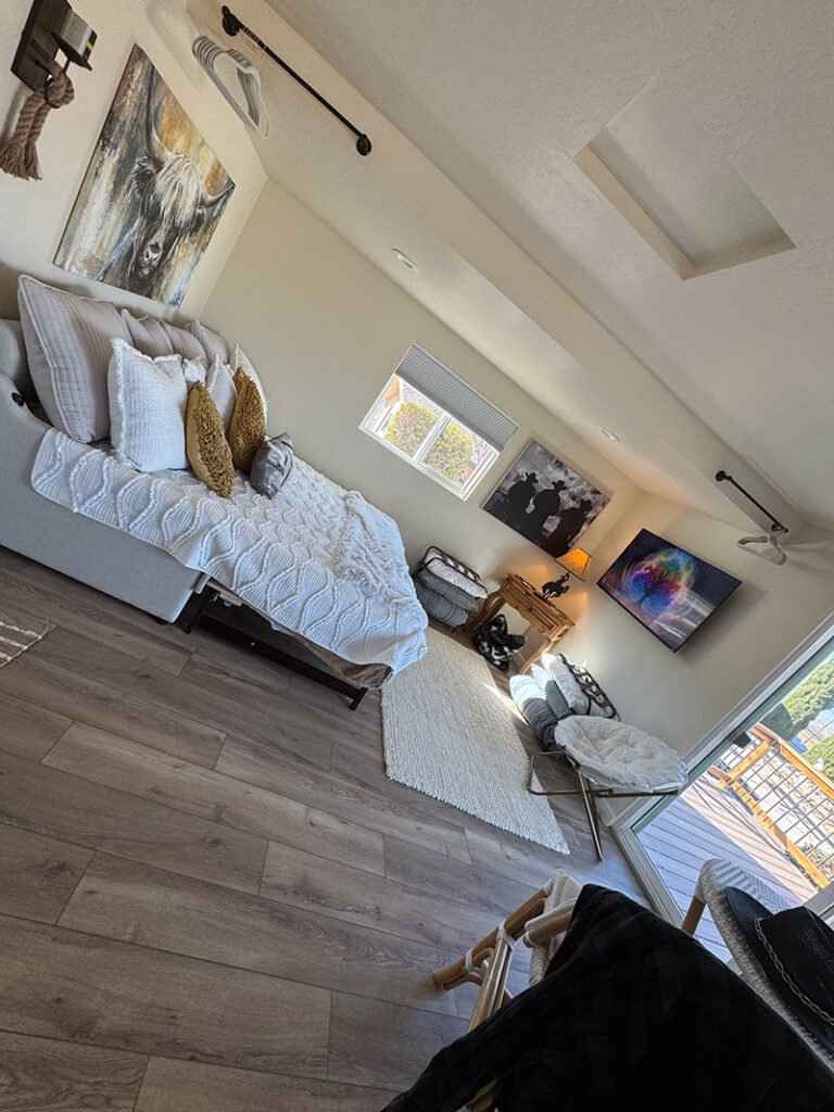 - Rustic Bedroom with Sitting Area A bedroom with white and mustard bedding, a highland cow painting, and wood plank flooring. A sitting area with cushioned chairs and wall art is near a sliding glass door leading to a deck. A coat rack and rope decor add character.